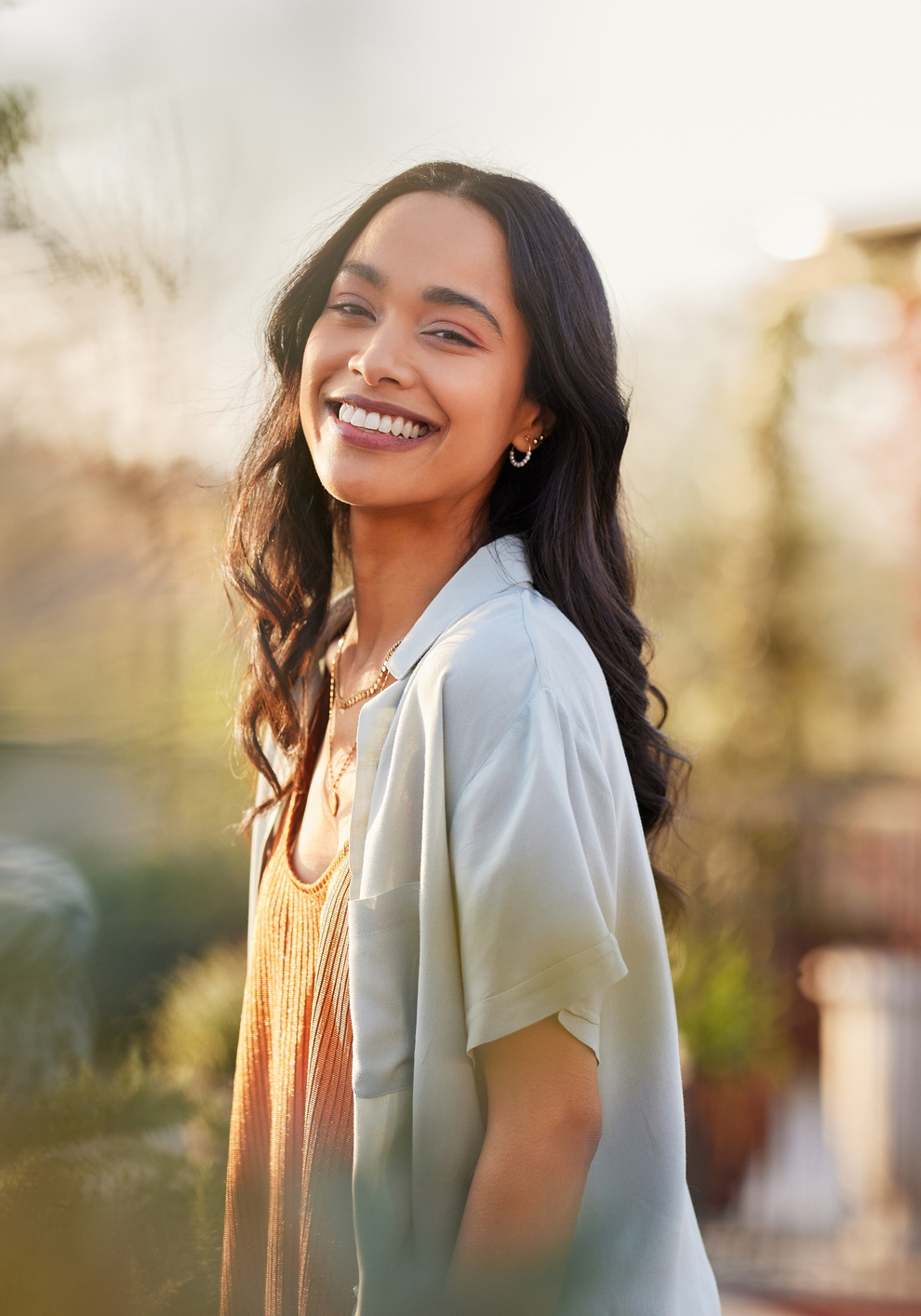Young latin woman in casual clothing in the garden looking at camera, during early morning. Portrait of healthy mexican girl enjoying nature during sunset. Mindful multiethnic woman enjoy morning ritual with fresh air.