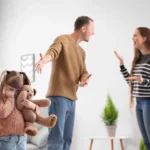 Parents arguing while the father gestures to their sad daughter holding a teddy bear, demonstrating co-parenting with a narcissist.