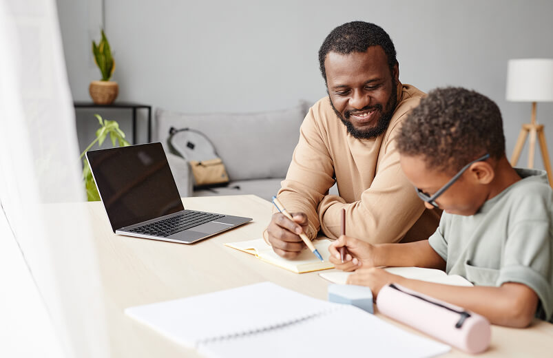 A smiling father helps his young son with homework.
