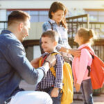 A smiling mother and father help their young son and daughter get ready outside a school building. The children wear backpacks, and the scene is filled with warm sunlight, suggesting a cheerful start to the school day.