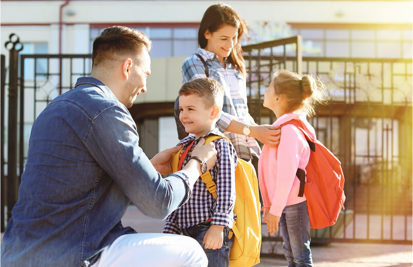 A smiling mother and father help their young son and daughter get ready outside a school building. The children wear backpacks, and the scene is filled with warm sunlight, suggesting a cheerful start to the school day.