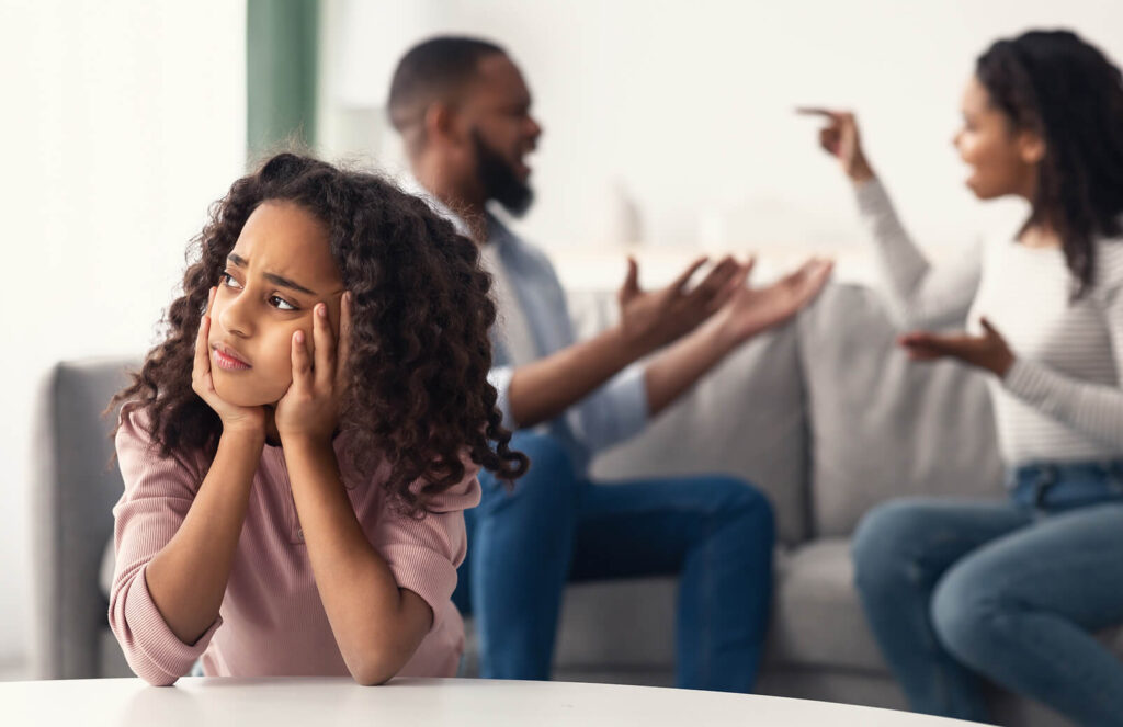 Upset child sitting in the foreground while adults argue on a couch in the background.