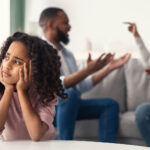 Upset child sitting in the foreground while adults argue on a couch in the background.