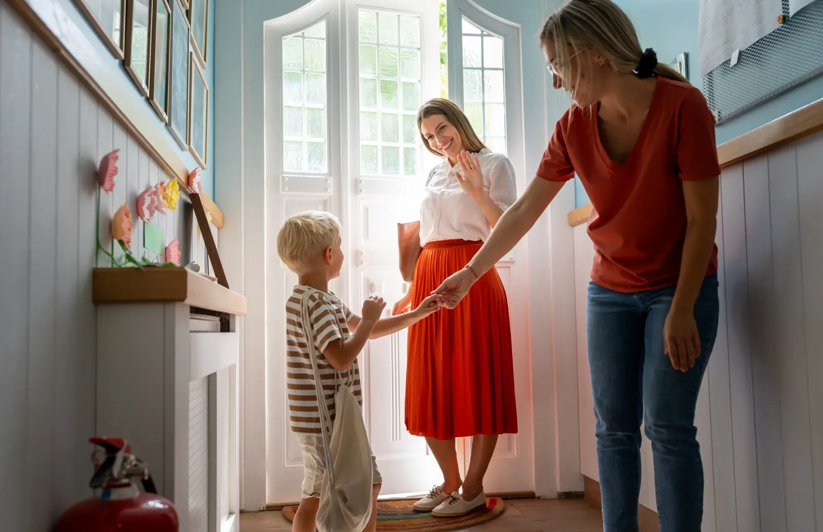 Young boy holding his mother's hand at the front door while another woman waves hello during a calm co-parenting transition