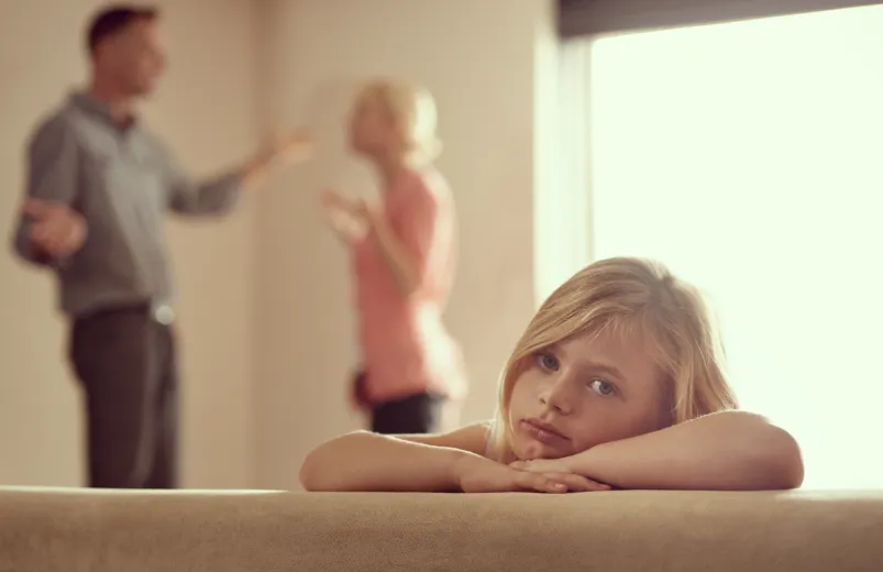 Sad child leaning on a couch with parents arguing in the background.
