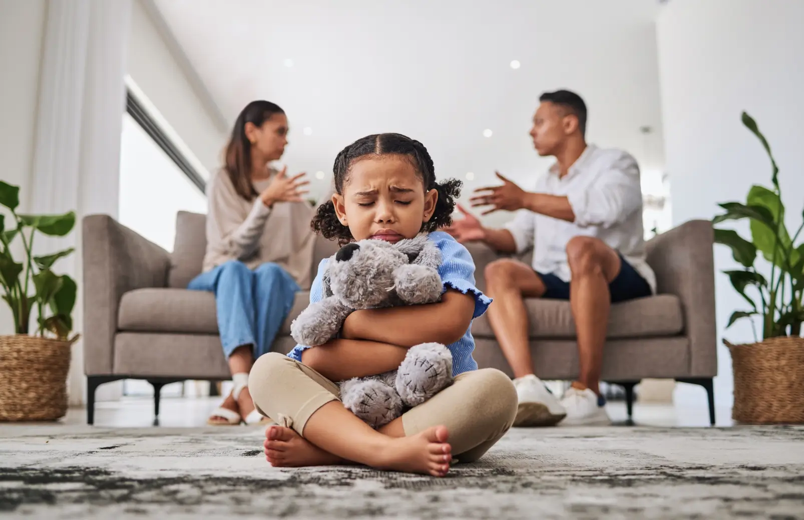 Young girl sitting on the floor hugging a stuffed animal with eyes closed while her parents argue on the couch behind her