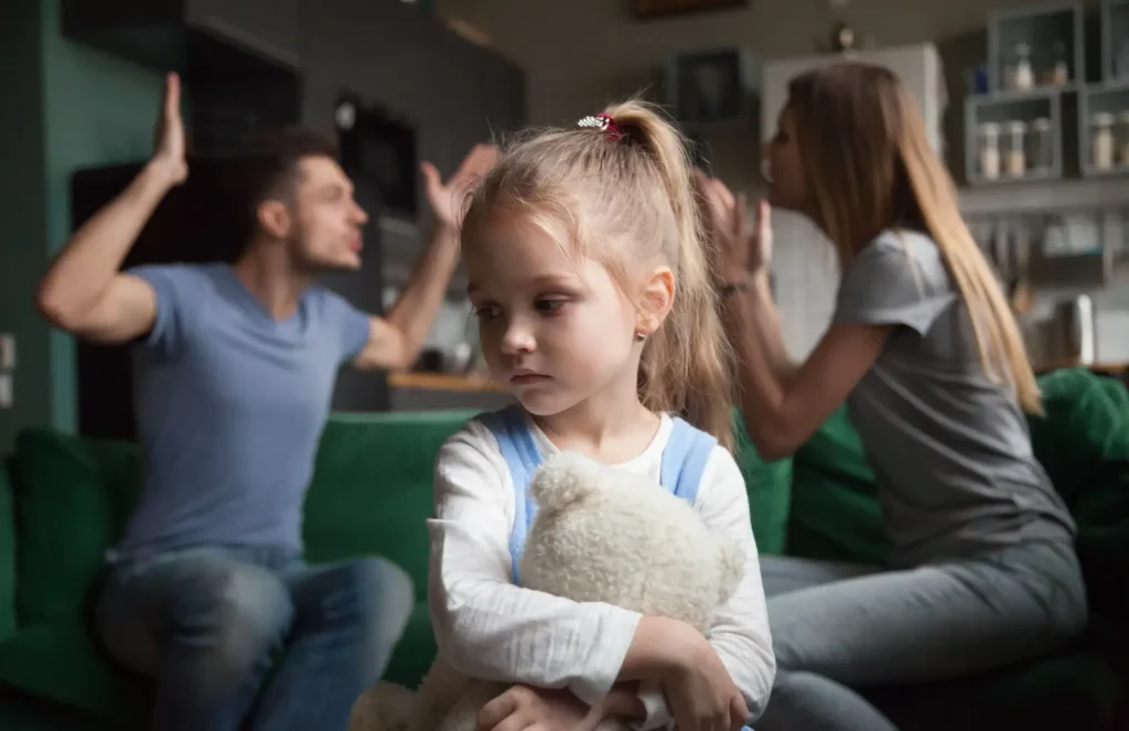 Young girl clutching a stuffed animal while her parents argue behind her on the couch