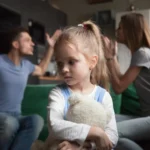 Young girl clutching a stuffed animal while her parents argue behind her on the couch