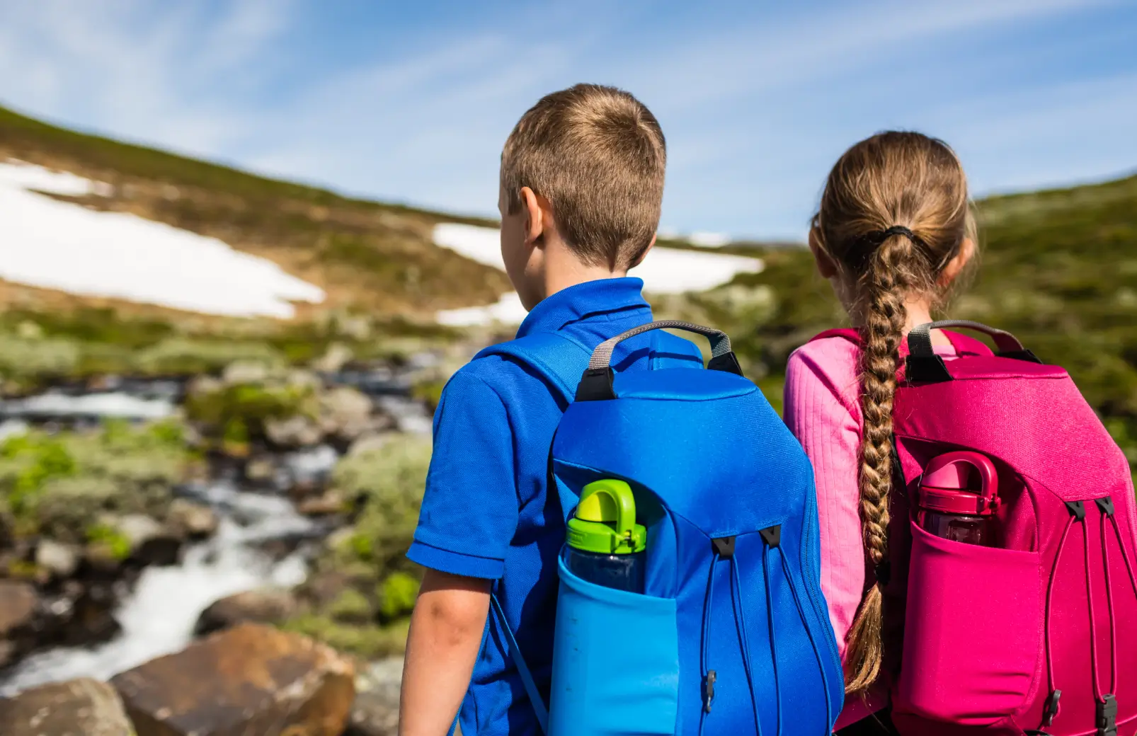 Two school-age children with backpacks looking out at a mountain stream during a summer adventure