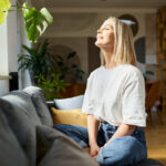 Happy adult woman sitting on the sofa with eyes closed enjoying bright daylight
