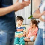 Two sad children sitting together at home while their parents argue, illustrating the signs a child may be struggling after a divorce